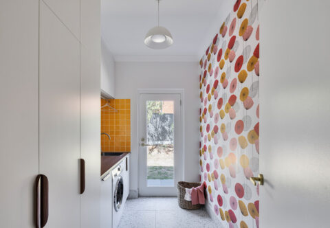 renovated laundry, mid-century style with yellow tiles and floral wallpaper, paired with white cabinetry and wood handles, designed with a deep sink, hanging rail, washer and glass door