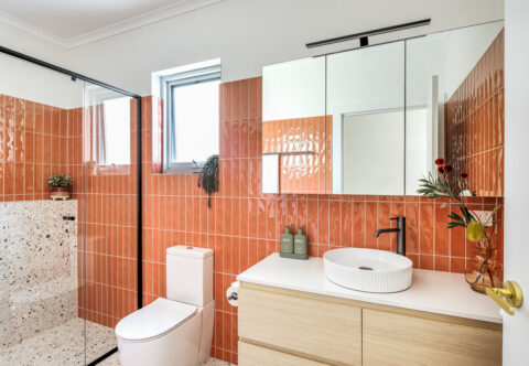 Renovated bathroom, mid-century style with orange wall tiles paired with terrazzo floor tiles, gunmetal fixtures and light wood finish cabinetry with white benchtop and fluted basin