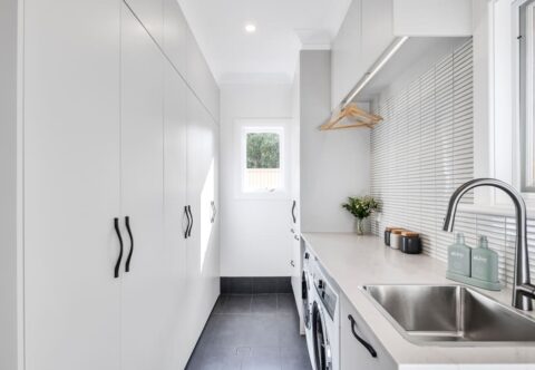 renovated laundry with kitkat feature tiles, custom cabinetry paired with black handles and dark grey floor tiles, new washer and dryer, clothes hanging rails and stainless-steel sink and tapware