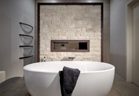 renovated bathroom, T-shaped dividing wall concealing shower and toilet, freestanding bath, textured split face travertine marble framed in veneer with nice in same material and LED strip lighting