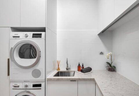 renovate laundry, minimal and functional with custom storage, stone benchtops, stacked washer and dryer, stainless steel sink and a discreet hanging rail.