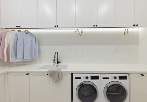 Renovated laundry with Hamptons style, warm white shaker cabinets, new washer and dryer, large benchtop with white sink and black fixtures and fittings, functional hanging rails for delicates.