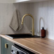 Renovated laundry with black sink, brushed brass tapware, tiled splashback, green cabinetry and laminated benchtops.