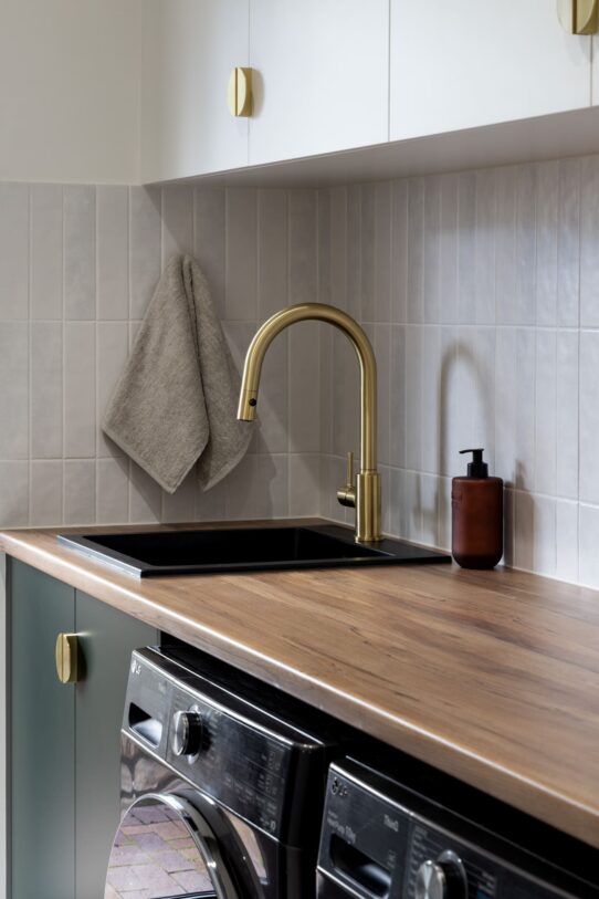 Renovated laundry with black sink, brushed brass tapware, tiled splashback, green cabinetry and laminated benchtops.